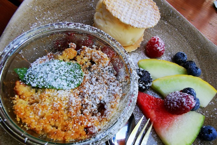 Dessert plate with crumble, ice cream sandwich, and assorted fruits on a brown plate.