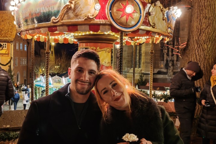 Smiling couple at festive carousel during nighttime, with others in the background.
