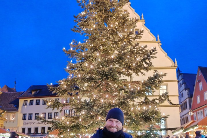Man in winter clothing smiling in front of a lit Christmas tree in a festive town square.