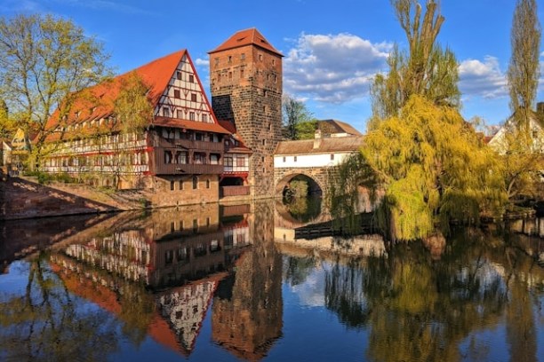 Historic buildings and tower reflected in calm river under blue sky.