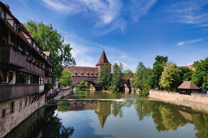 Medieval-style buildings with river and bridge, trees, and blue sky.