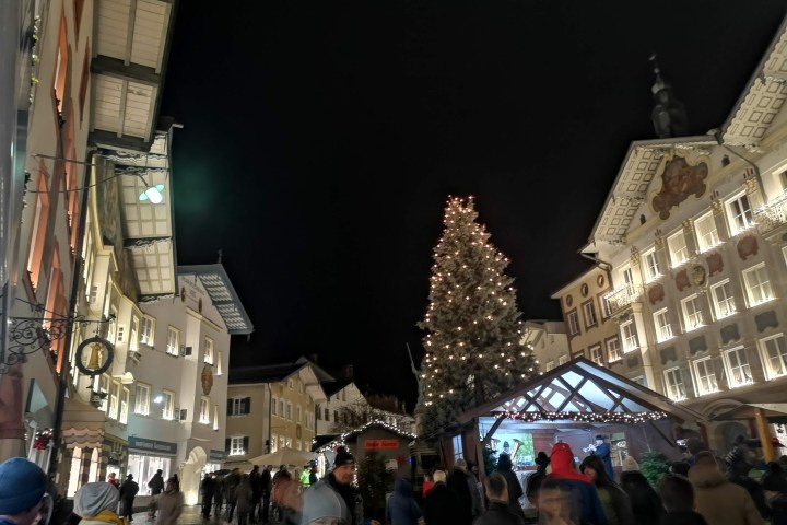 Festive night market with Christmas tree and lights, surrounded by people and illuminated buildings.