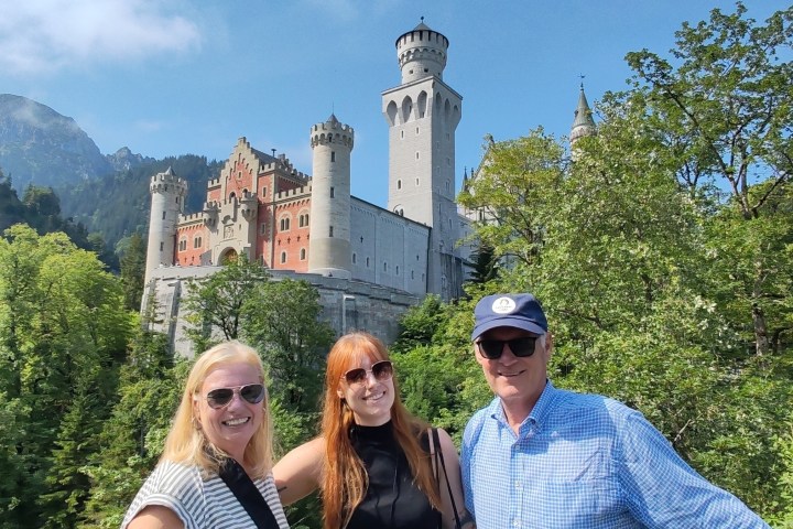Three people smiling in front of a castle surrounded by trees on a sunny day.