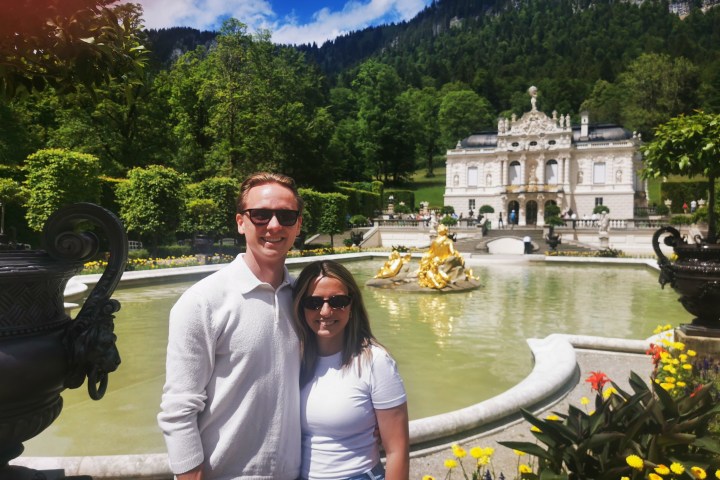 Couple posing in front of a fountain with a historic building and mountains in the background.