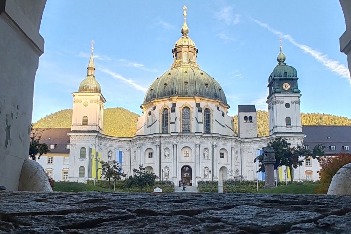 Majestic dome of a baroque church framed by an archway, with mountains and a clear sky in the background.