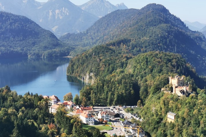 Scenic view of a castle surrounded by forested hills and a lake with mountains in the background.