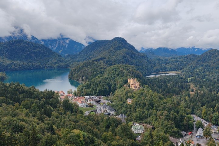 Scenic view of a castle by a lake surrounded by mountains and forests under a cloudy sky.