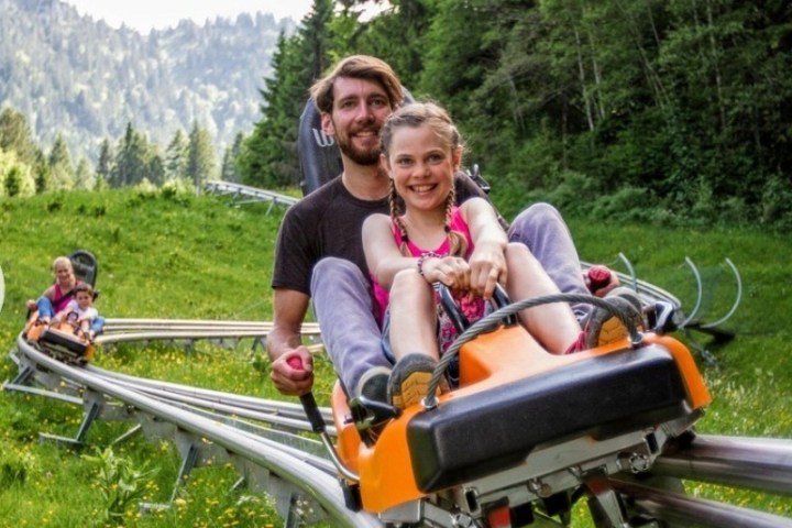 Man and child riding an orange alpine coaster through a forested area.
