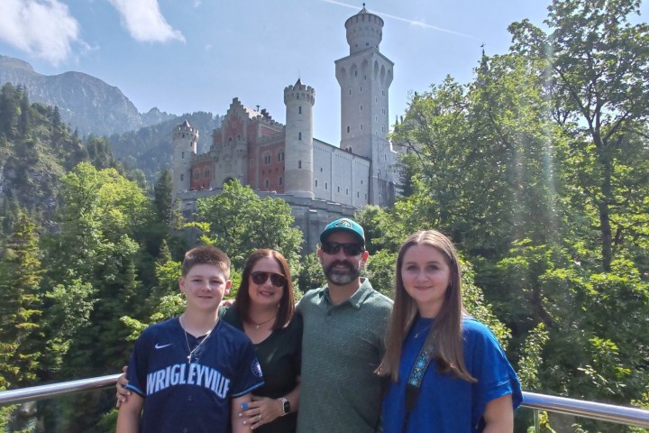 Family of four posing in front of a castle surrounded by trees and mountains.