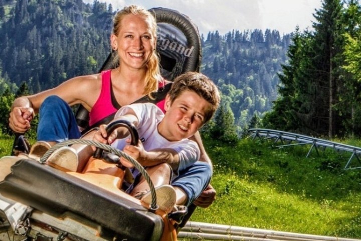 Smiling woman and boy on an alpine coaster ride with forested mountains in background.