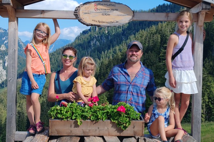 Family posing under wooden structure with logs and flowers, mountains in background.