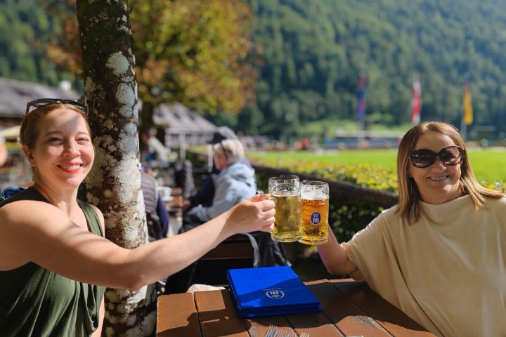 Two women clinking beer mugs at an outdoor table with a scenic mountain backdrop.