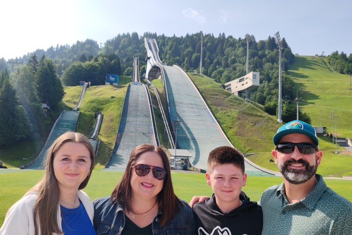 A family poses in front of a large ski jump with green forested hills.