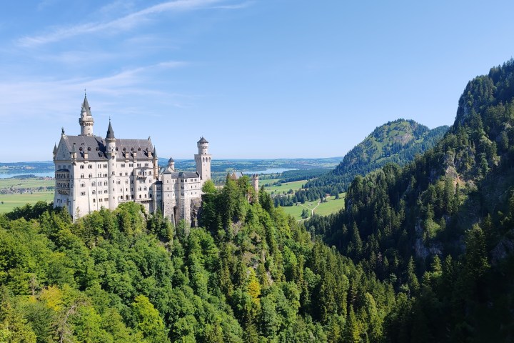 Neuschwanstein Castle surrounded by trees with mountains in the background on a clear day.