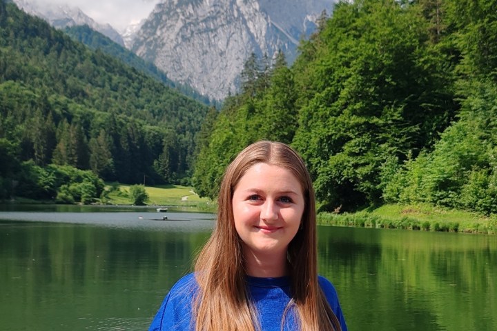 Person in blue shirt smiling in front of a lake and green forest with mountains in the background.