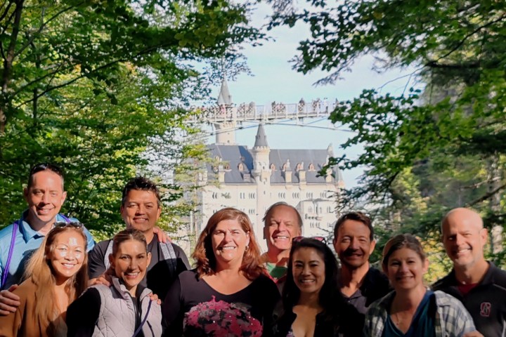 Group of people posing with Neuschwanstein Castle in the background among trees.