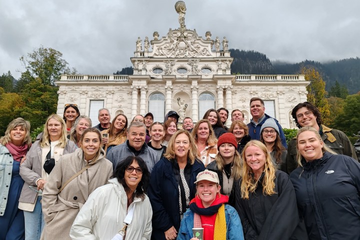 Group of 27 people smiling in front of an ornate building with cloudy skies and greenery.
