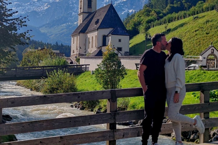 Couple kissing on bridge with church, river, and snowy mountains in background.