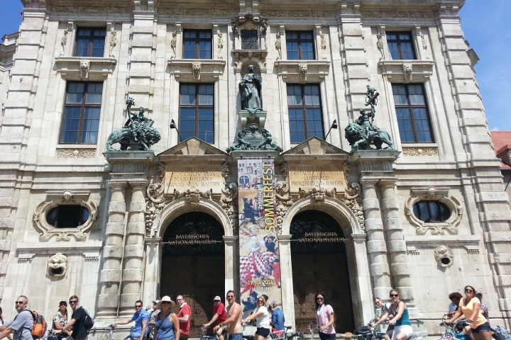 People on bicycles in front of a historic building with decorative statues.