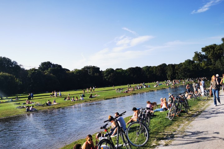 A park scene with people relaxing by a small river, bicycles along the path, and trees in the background.
