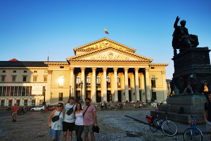 Four people posing in front of a neoclassical building with columns and a statue at sunset.