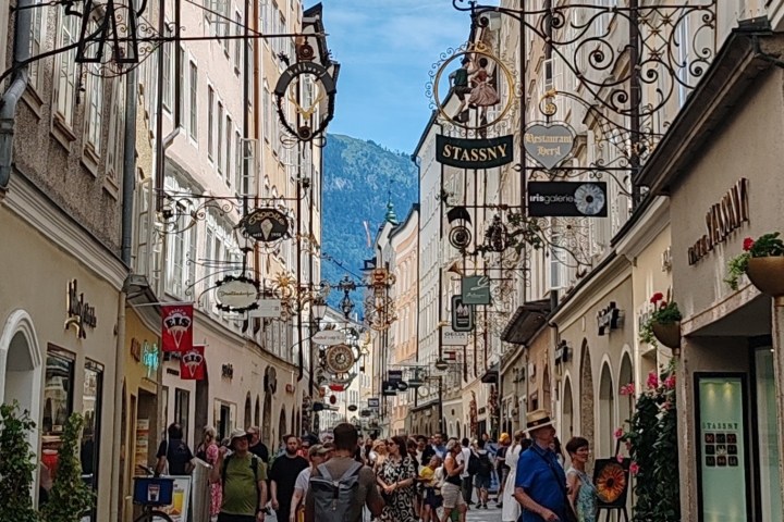 Narrow street in a European city with people, shops, and decorative signs.