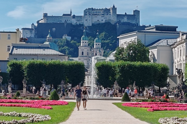 Colorful garden with path and fountain, castle on hill in background, sunny day.