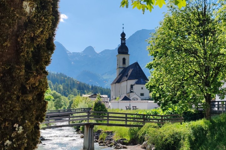 Scenic view of a church near a wooden bridge over a stream, surrounded by trees and mountains.