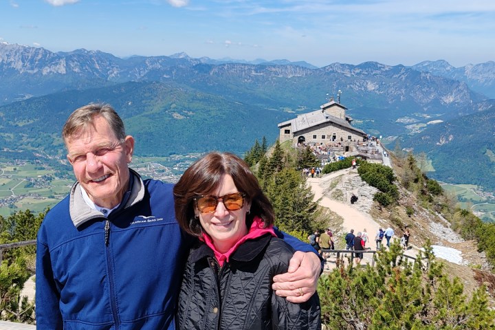 Two people smiling on a mountain path with a scenic view of a chalet and mountains in the background.