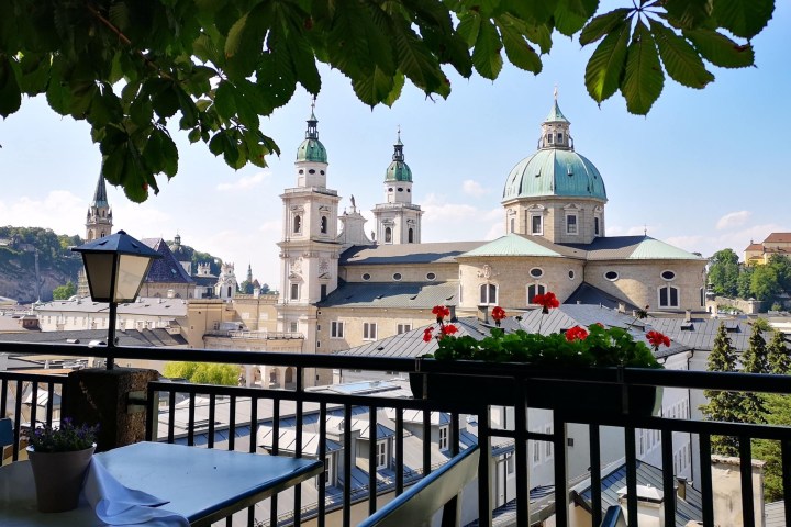 View of Salzburg Cathedral with table, chairs, and flowers on a shaded terrace.