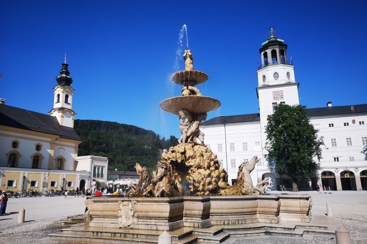 Ornate fountain in a plaza with historical buildings and a clear blue sky.