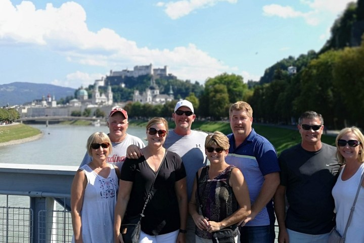 A group of seven people posing on a bridge with a scenic city and hills in the background.