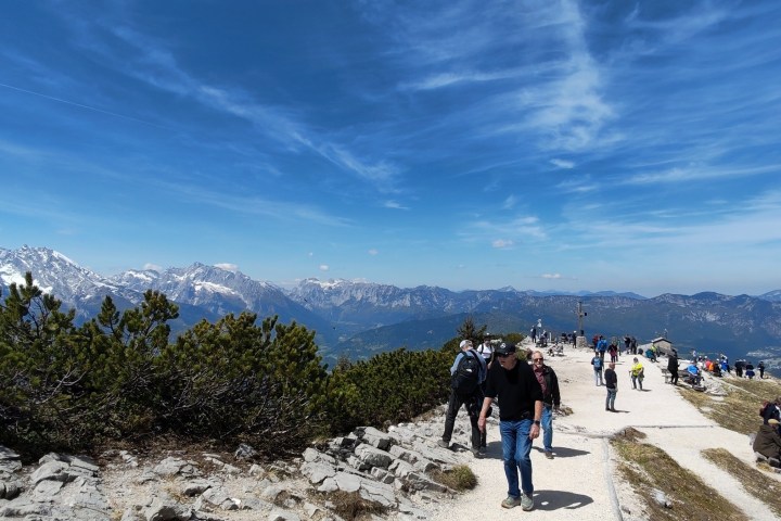 People walking on a mountain path with a scenic view of distant snow-capped peaks under blue sky.