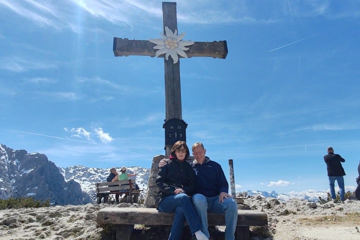 Couple sitting by mountain cross with scenic view and benches around.