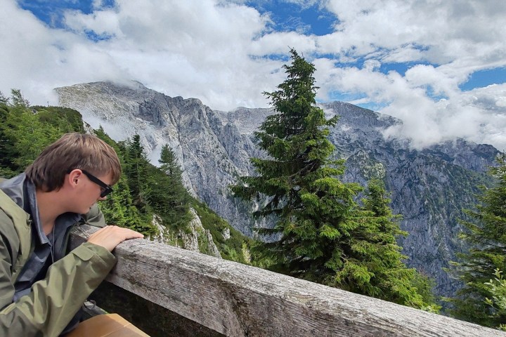 Person in a jacket leans on a wooden railing, overlooking a mountain landscape with trees and clouds.