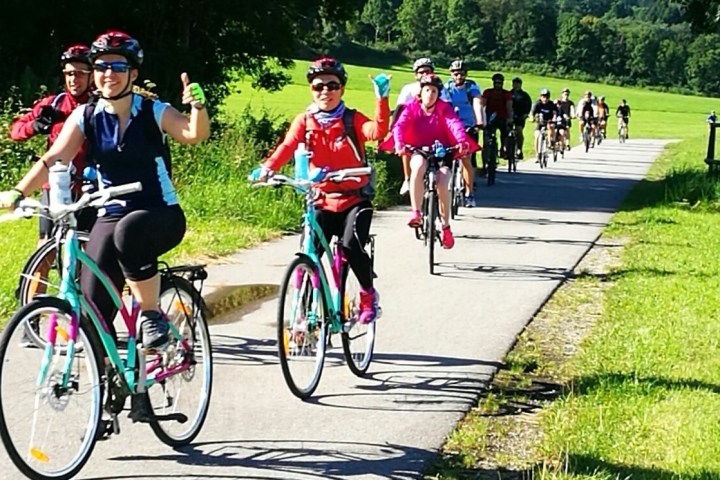 Cyclists riding on a path through a sunny green landscape, some giving thumbs up.