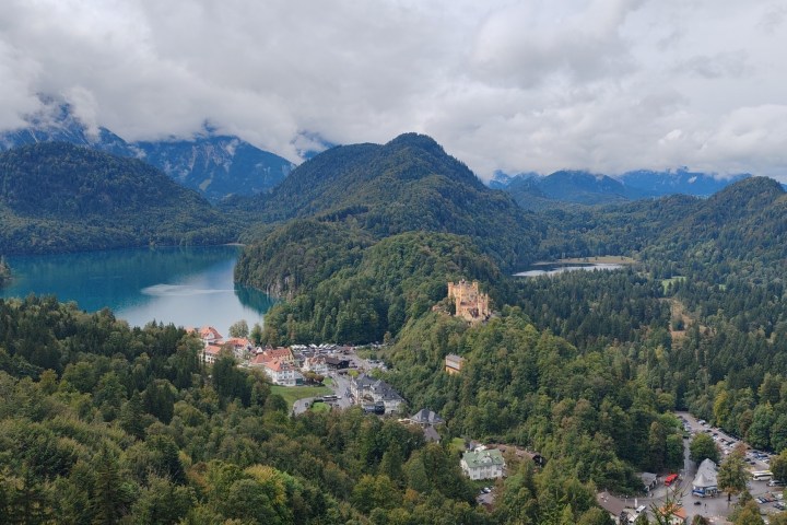 Scenic view of a castle on a hill surrounded by forests, lakes, and mountains on a cloudy day.