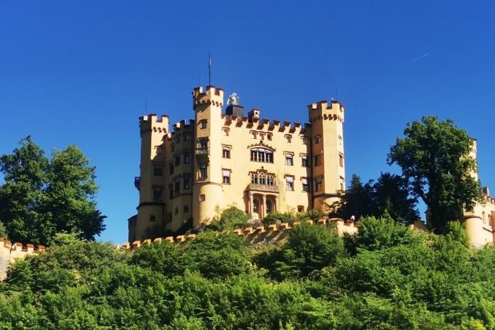 Yellow castle with turrets on a hill surrounded by trees against blue sky.