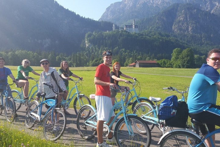 Group of people on bicycles in a scenic landscape with mountains and a castle in the background.