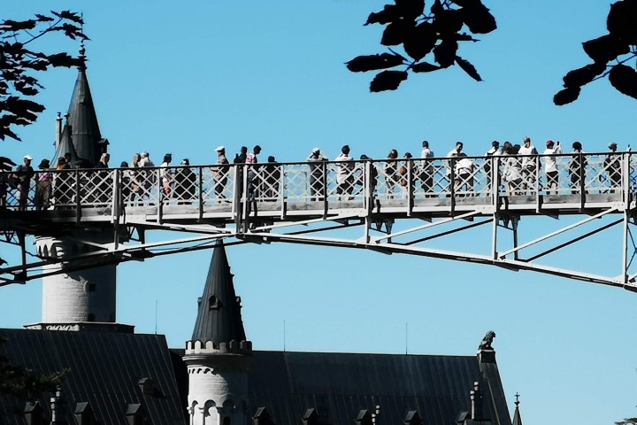 People on a bridge above a castle with pointed towers and a clear blue sky.
