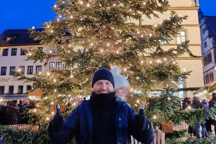 Person smiling in front of a lit Christmas tree at a festive market in the evening.
