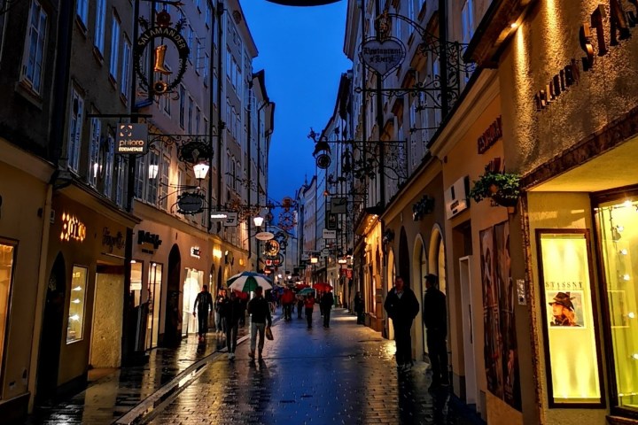 Narrow cobblestone street at dusk with lit shop windows and people walking under umbrellas.