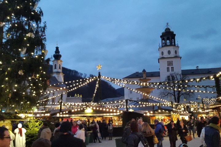 People at a festive outdoor market with string lights and decorated buildings in the evening.
