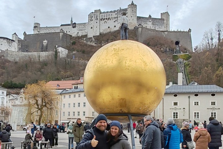 Couple posing in front of large golden sphere sculpture with a statue on top, fortress in background.