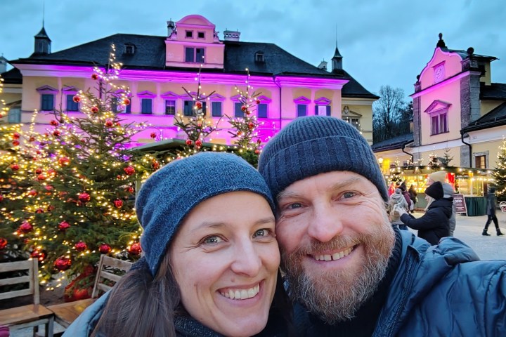 Smiling couple in winter attire at festive market with lit trees and pink-lit building in background.