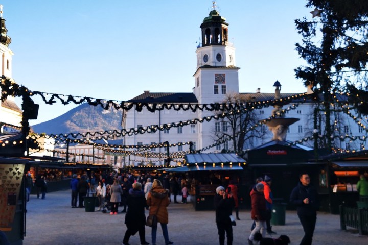 People walking through a festive outdoor market with string lights and a clock tower in the background.