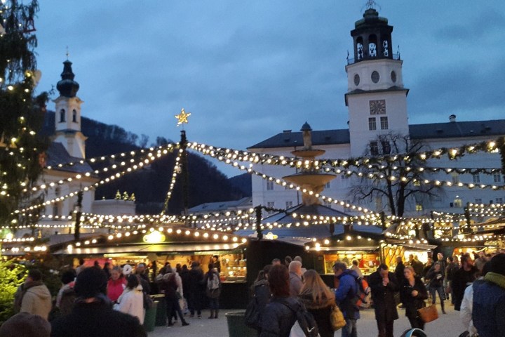 Crowded Christmas market with lights, stalls, and a large tree in the foreground under an evening sky.
