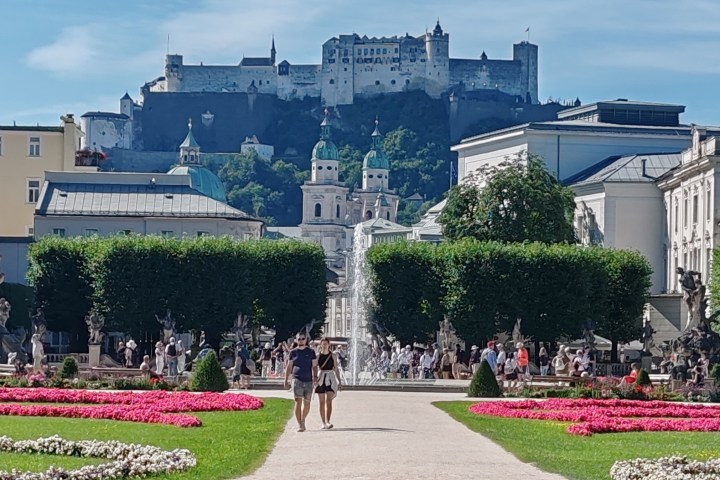 Castle on a hill overlooking a busy garden with people, trees, and pink flowers.
