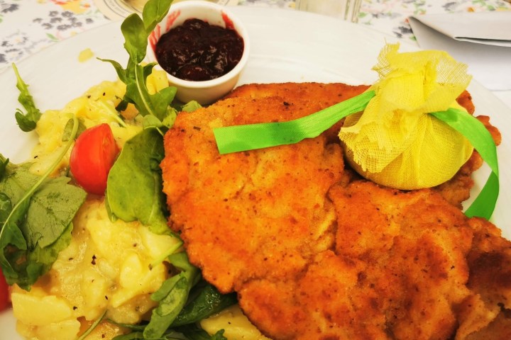 Breaded schnitzel with potatoes, arugula, cherry tomatoes, jam, and a lemon wrapped in yellow cloth on a plate.