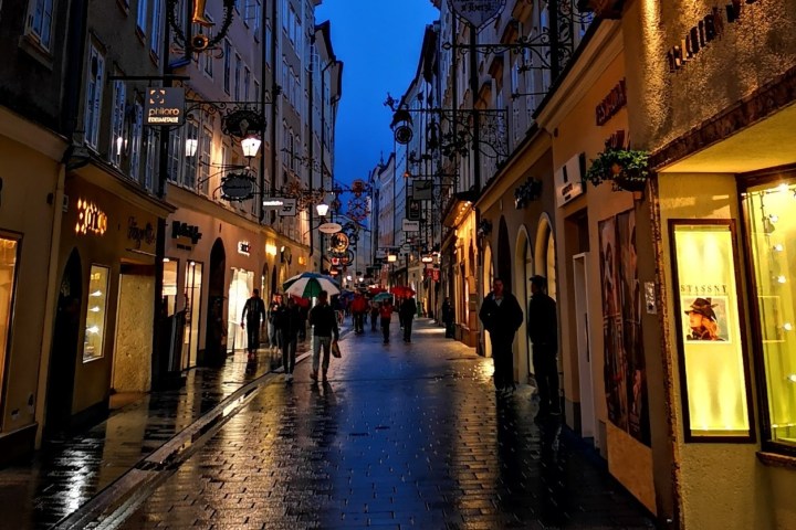 People walking with umbrellas on a narrow, wet street with lit shop windows at dusk.